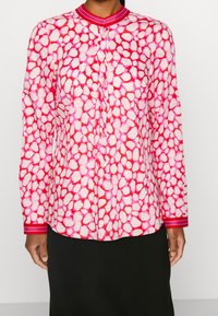 Camicia a maniche lunghe di colore rosa con un motivo astratto di forme bianche irregolari, dotata di un colletto e polsi solidi rosa con accenti rigati.
