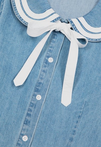 Camicia in denim azzurro chiaro con bottoni bianchi e un ampio colletto da marinaio bianco con righe blu, legato con un fiocco bianco sul collo.
