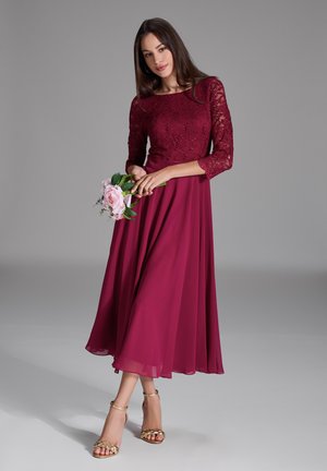 Robe en dentelle bourgogne avec des manches trois-quarts ; jupe en chiffon fluide ; modèle tenant un bouquet de fleurs roses ; sandales dorées à lanières.
