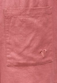 Κοντινό πλάνο ενός ξεθωριασμένου ροζ υφασμάτινου τσέπης με ένα κεντημένο λογότυπο σε σχήμα καρδιάς στην κάτω δεξιά γωνία.