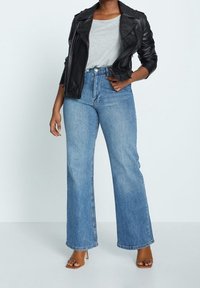 Zwarte leren motorjack over een grijs T-shirt, gecombineerd met lichtblauwe wijde jeans en bruine sandalen met hakken, wat een casual outfit laat zien.