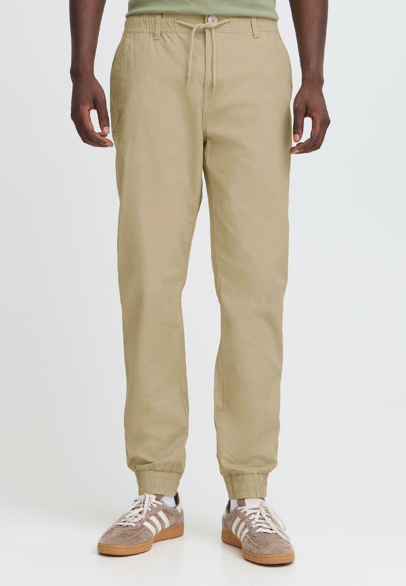 Pantalons de jogging beiges avec une taille élastique et un cordon de serrage. Poignets ajustés aux chevilles. Portés avec des chaussures de sport grises et blanches.