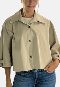 Beżowa krótka koszula z dużym kołnierzem, zapięciem na guziki i podwijanymi rękawami, wykonana z gładkiej tkaniny, zestawiona z niebieskimi dżinsami.