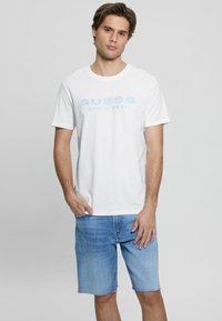 Witte katoenen T-shirt met lichte blauwe opdruk "GUESS SINCE 1981", gecombineerd met lichte blauwe denim shorts. Dit outfit heeft een casual pasvorm en korte mouwen.