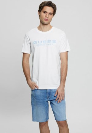 T-shirt en coton blanc avec impression "GUESS SINCE 1981" bleu clair, associé à un short en denim bleu clair, présentant une coupe décontractée et des manches courtes.