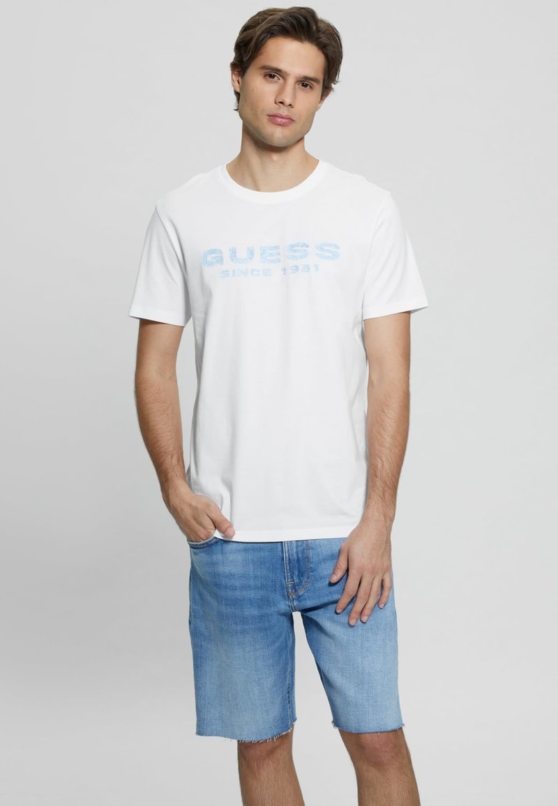 Witte katoenen T-shirt met lichte blauwe opdruk "GUESS SINCE 1981", gecombineerd met lichte blauwe denim shorts. Dit outfit heeft een casual pasvorm en korte mouwen.