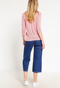 Kvinde med langt blondt hår iført en pink bluse med trekvartærmer, korte blå jeans og hvide sneakers, stående med ryggen til.