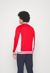 Rød jakke med hvite sidelapper, stripete krage og mansjetter. Laget av glatt stoff, slimmet design og lange ermer.