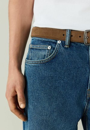 Left hand resting beside blue denim jeans with a brown suede belt and white tucked-in shirt.