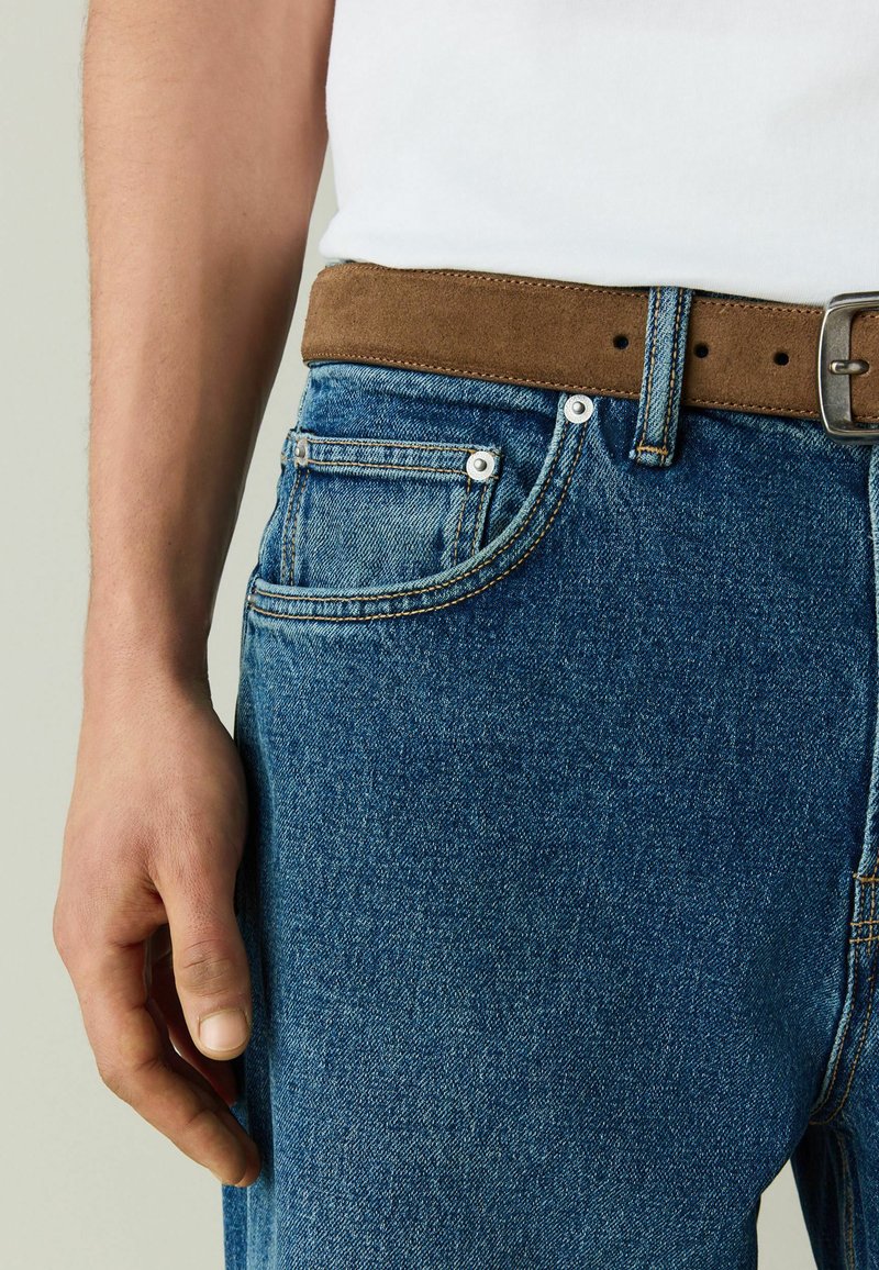 Main gauche posée à côté d'un jean en denim bleu avec une ceinture en suède marron et une chemise blanche rentrée.