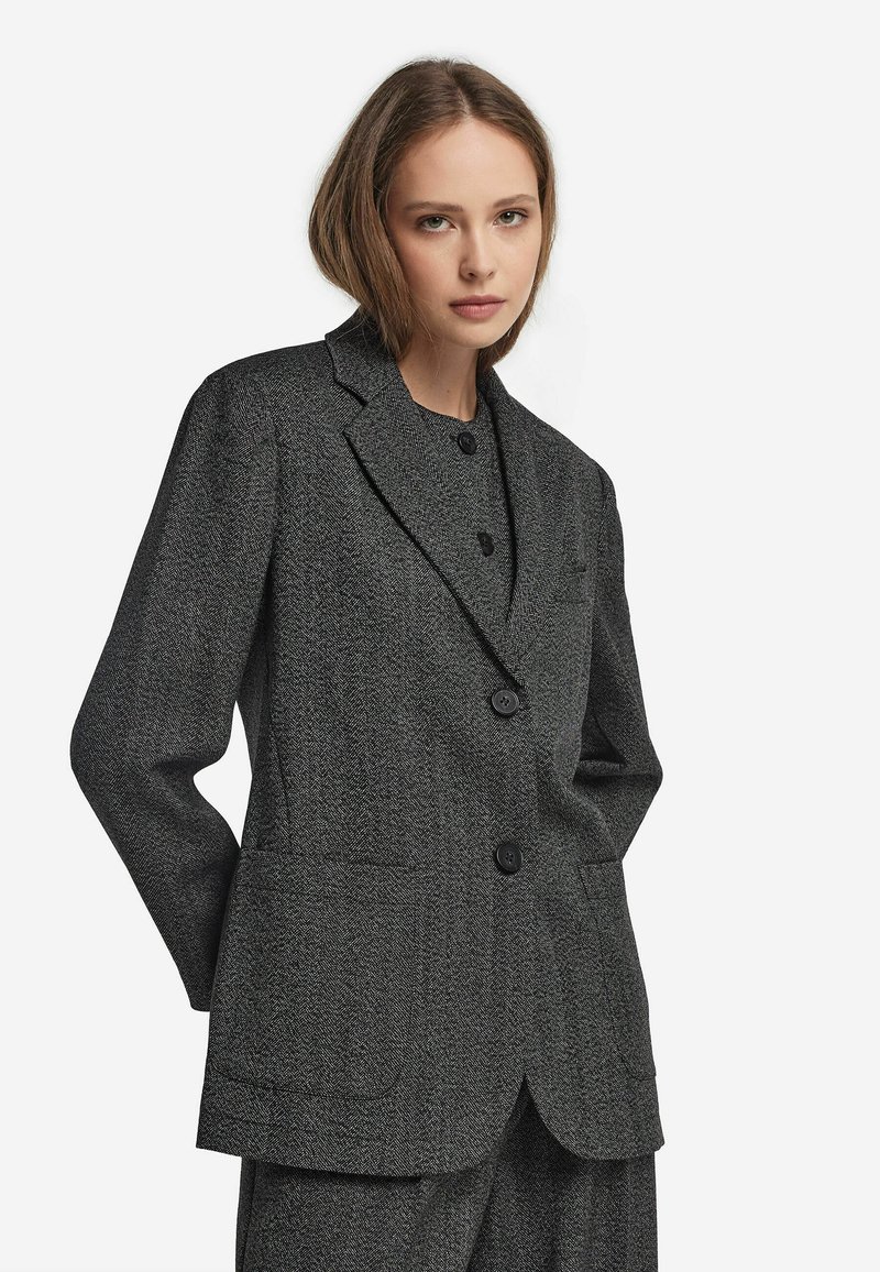 Ung kvinna i en mörkgrå herringbone-blazer och matchande byxor, stående mot en enkel vit bakgrund.