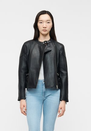 Femme portant une veste en cuir noire asymétrique sur un haut blanc et un jean bleu clair, debout devant un fond uni.