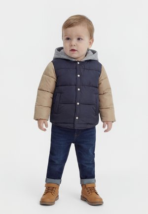 Νήπιο όρθιο, φοράει μια καπιτονέ ναυτική και καφέ ζακέτα με γκρι κουκούλα, σκούρα τζιν γυρισμένα μπατζάκια και καφέ δερμάτινα μποτάκια σε λευκό φόντο.