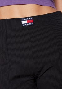 Černé žebrované džíny s páskem, na kterém je obdélníková značka Tommy Jeans v námořnické modré, červené a bílé barvě. Texturovaná látka s jemnými vertikálními pruhy.