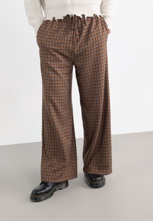 Person trägt braune, karierte, weit geschnittene Hose mit elastischem Bund und schwarze Stiefel mit dicker Sohle, steht auf grauem und weißem Boden.