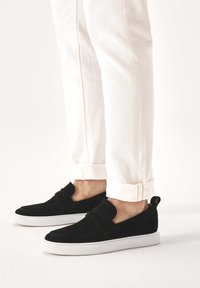 Zapatos negros de ante sin cordones con suela de goma blanca, con un detalle decorativo de tira y puntera redondeada, combinados con vaqueros blancos remangados.
