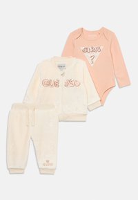 Három részes baba öltözet, beleértve egy barack színű, hosszú ujjú bodysuitot logóval, egy krém színű, cipzáros dzsekit flitteres "GUESS" felirattal, és egy hozzá illő krém színű nadrágot.