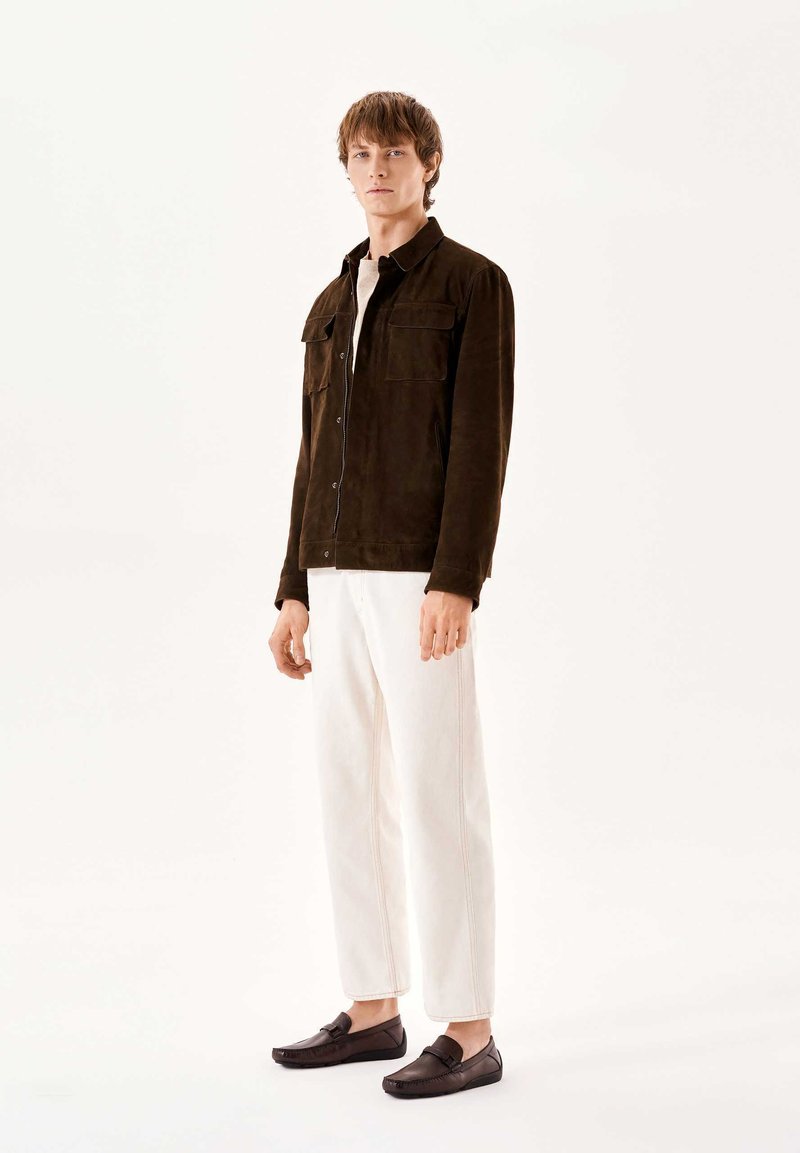 Bruine suede overhemdjasje met twee borstzakken, een witte t-shirt eronder, crèmekleurige tapse broek en donkere loafers, staande op een witte achtergrond.