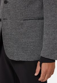 Gray herringbone blazer with notched lapels, two buttons, and a pocket. Features a contrasting black inner cuff with a thumb loop.