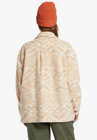 Fleecejack in crème met een textuur van geometrische patronen, voorzien van een kraag, lange mouwen en een losse pasvorm, gecombineerd met een oranje gebreide beanie.