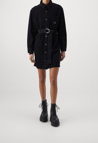 Persoon in een zwarte denim jurk met ceintuur en zwarte veterschoenen met platform, staande tegen een effen lichte achtergrond.