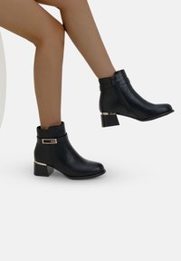 Botas de tobillo negras con un acabado de cuero suave, tacón grueso y un acento metálico decorativo. Presentan una cremallera lateral para facilitar el uso.