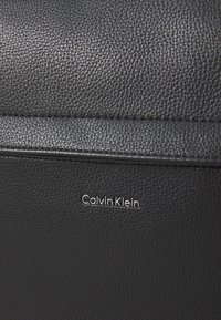 Textura de cuero negro con grano visible, que presenta un sutil logotipo en relieve de "Calvin Klein" en tono plateado. Diseño simple y moderno.