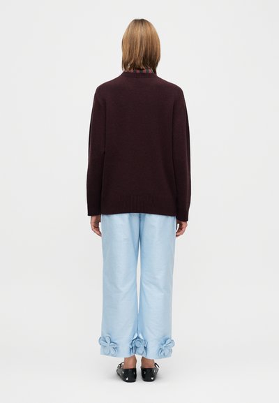 Mørkerød genser med avslappet passform og strikket struktur. Lyseblå bukser med blomsterdetaljer ved mansjettene. Sorte flate sko. Nøytral bakgrunn.