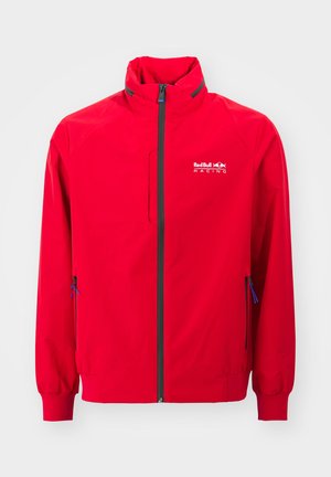 Červená větrovka v živé červené barvě, s vysokým límcem, černým zipem, kapsou na hrudi a modrými akcenty na zipu. Zobrazena ikona Red Bull Racing.