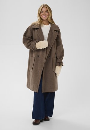 Hnědý vlněný kabát s velkými klopami a dvěma předními kapsami, doplněný krémovými rukavicemi a modrými širokými džíny. Světlé pozadí.