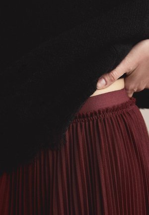 Burgunderroter Faltenrock mit elastischem Bund, kombiniert mit einem schwarzen, strukturierten Oberteil, eine Hand passt den Bund an.