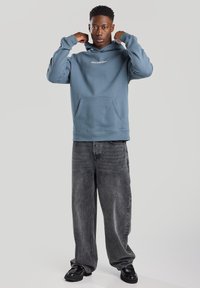 Blue cotton hoodie with front pocket and "Open Your Mind" embroidered text. Paired with faded black wide-leg jeans and black shoes.