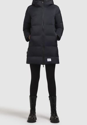 Černý prošívaný kabát po kolena se zapínáním na zip vpředu, bočními kapsami a kapucí, doplněný černými legínami a černými vojenskými botami na šněrování.