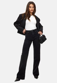 Zwarte denim wijdpijpenbroek gecombineerd met een aansluitend wit tanktop en een oversized zwarte blazer. Houdt een kleine zwarte handtas vast.