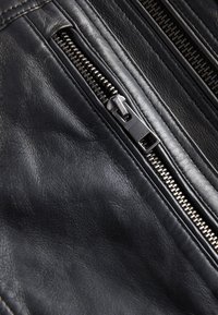 Gros plan sur un cuir noir avec une fermeture éclair métallique argentée partiellement ouverte, mettant en valeur la texture et les détails des coutures.