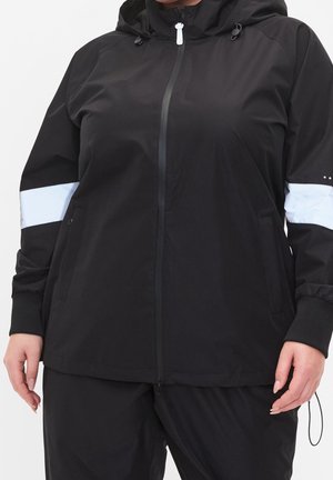 Sort lynlåset hættejakke med hvide refleksbånd på overarmene, båret af en person med hænderne afslappet langs siden.