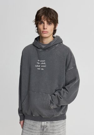 Felpa grigia con finitura testurizzata, con tasca frontale e testo bianco. Taglio oversize con cappuccio con coulisse e polsini a costine.