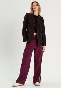 Blazer noir au design ajusté, associé à un pantalon large bordeaux avec des rayures sur les côtés. Le modèle porte des chaussures à enfiler de couleur claire.