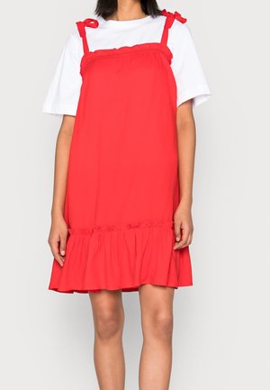 Woman wearing a sleeveless red dress with bow-tied straps over a white short-sleeve T-shirt, standing against a plain light background.