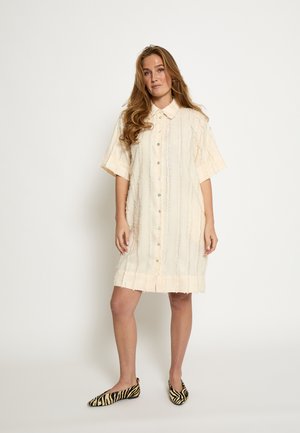 Robe à manches courtes boutonnée, de couleur crème clair avec des rayures verticales texturées et des bords effilochés. Associée à des ballerines rayées noires et blanches.