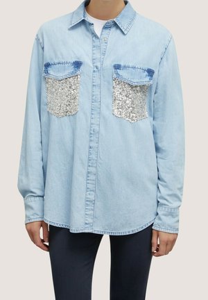 Camisa de mezclilla azul claro con cierre de botones al frente, mangas largas, dos bolsillos decorados con lentejuelas y un corte holgado. Textura suave, diseño informal.