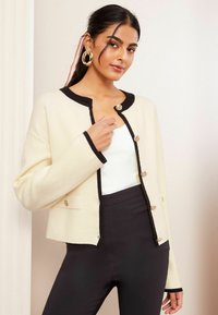 Cardigan crem cu detalii negre, cu guler rotund și butoane aurii. Stylistic purtat peste un top alb și asortat cu pantaloni negri.