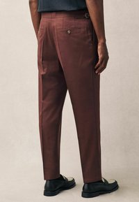 Brown tailored trousers made from a smooth fabric, featuring two back pockets with button closures, belt loops, and a slim fit design.