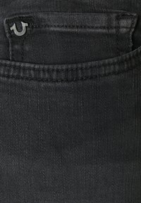 Černé džíny z denimu mají náprsní kapsu s logem. Tkanina má hladkou strukturu s minimálním vyblednutím a jemnými švy.