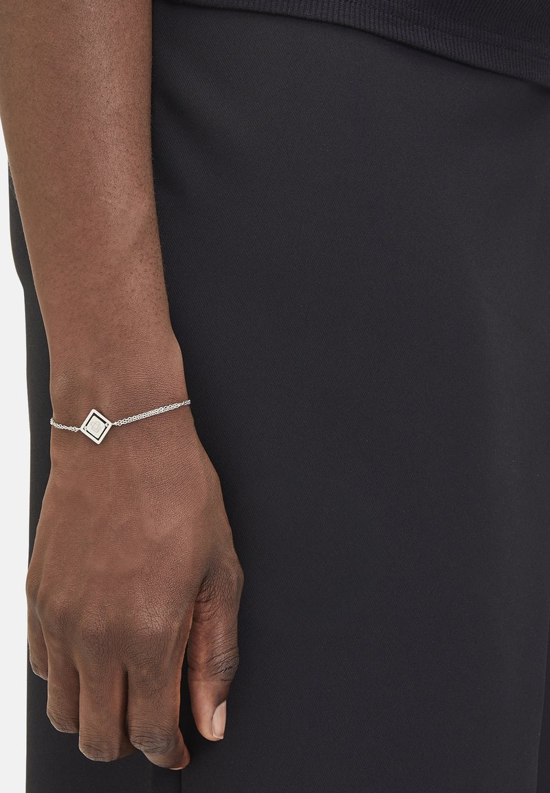 Silbernes Armband mit charm in quadratischer Form, zarter Kette und strukturierten Oberfläche, getragen an einem dunkel gekleideten Handgelenk in Nahaufnahme.