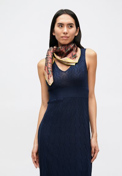 Hodvábna šatka s geometrickým cikcakovým vzorom v červenej, hnedej a žltej farbe na krémovom pozadí, ohraničená žltým lemom.