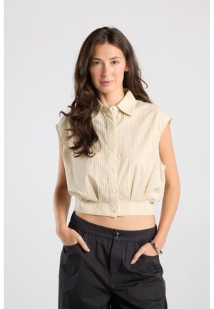 Jonge vrouw met lang donker haar die een mouwloos beige cropped blouse met knopen en zwarte broek draagt met handen in de zakken.