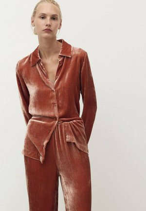 Il set di camicia e pantaloni in velluto ruggine presenta un colletto, un'apertura frontale con bottoni e una vita elastica con un dettaglio annodato. Tessuto morbido, tonalità arancione calda.