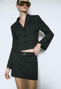Sort blazer med stikninger og tætsiddende design, to knapper og udvidede ærmer, parret med en matchende mini-skirt og bælte-detalje.