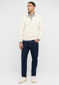 Jasno beżowy sweter z zamkiem przy kołnierzu, narzucony na jasnoniebieską koszulę. Ciemnoniebieskie dżinsy i białe trampki dopełniają strój.